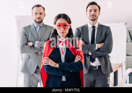 super businesswoman standing with crossed arms in mask and cape with businessmen in office Stock Photo