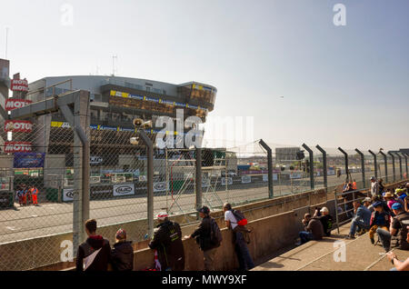 19-20th May 2018. Le Mans, France.  Behind the scenes at the MotoGP.  Spectators overlooking the race circuit's start finish line and the back of the grid.  The official's grand stand is opposite behind the entrance to pit lane.  A tannoy address system is mounted on top of the perimeter fencing.  It's a bright sunny day with a blue sky and the sun behind the grand stands opposite. Stock Photo