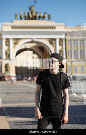 Young stylish man tourist walking near General Staff Builsing arch in the palace square in St. Petersburg. He wearing in black jeans, black T-shirt, black cap with riund sunglasses. Stock Photo