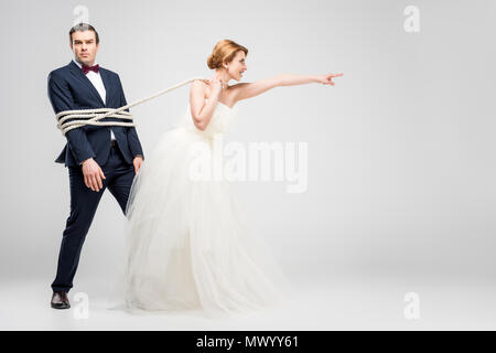 bride pointing and pulling groom bound with rope, isolated on grey, feminism concept Stock Photo