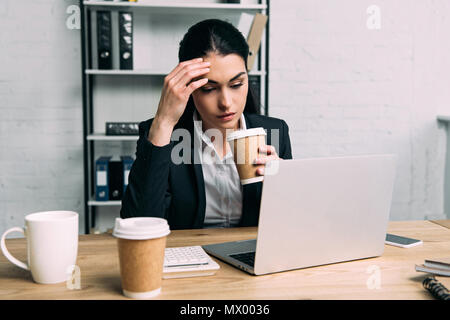 tired businesswoman in suit with coffee to go working on laptop at workplace in office Stock Photo