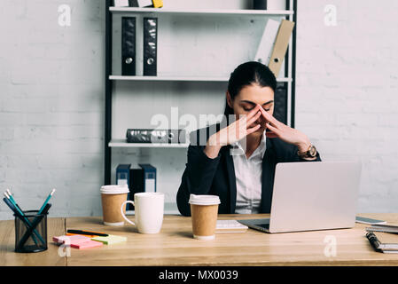 portrait of overworked businesswoman in suit at workplace with laptop and coffee to go in office Stock Photo