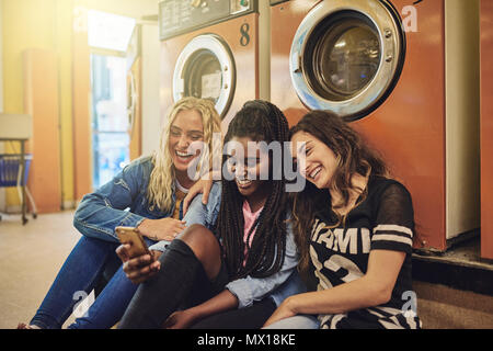 Group of young female friends sitting on a laundromat floor together laughing and using a cellphone Stock Photo