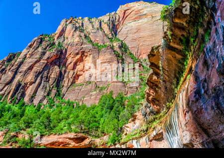 Water running off rocky ledge and  tall reddish mountain with green trees in background under blue sky. Stock Photo