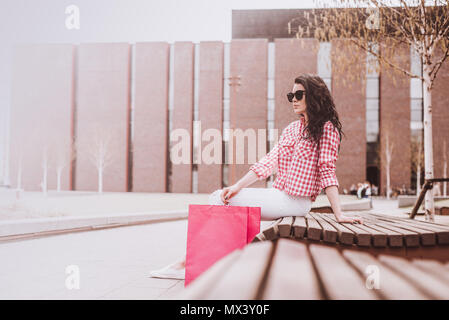 A woman resting after shopping. Attractive girl sitting on a bench holding shopping bags next to. Subdued and toned colors. Vintage and retro styles.  Stock Photo