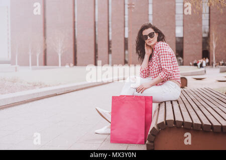 A woman resting after shopping. Attractive girl sitting on a bench holding shopping bags next to. Subdued and toned colors. Vintage and retro styles.  Stock Photo