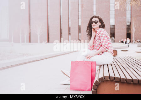 A woman resting after shopping. Attractive and cheerful girl sitting on a bench holding shopping bags next to it. Subdued and toned colors. Vintage an Stock Photo