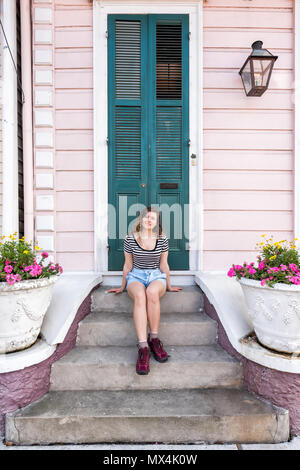 Young hipster millennial woman sitting smiling happy on stairs steps, front porch in New Orleans by colorful pink door architecture during day with ye Stock Photo