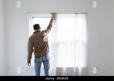 Young man standing in front of bright window in room adjusting blinds curtains, white painted walls, during remodeling, cleaning, inspection Stock Photo