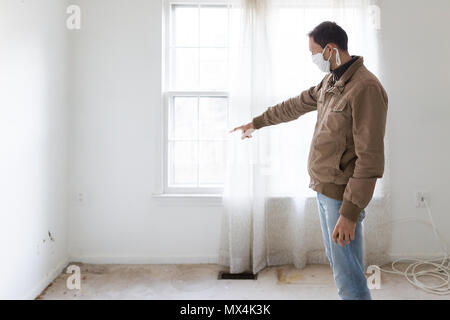 Young man in mask standing pointing at room wall carpet floor flooring, white painted walls, during remodeling renovation, cleaning, inspection of dir Stock Photo