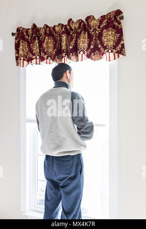 Young man standing in front of bright window in room adjusting old-fashioned retro blinds curtains Stock Photo