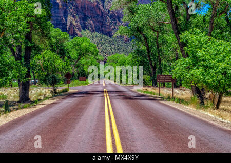 Looking down the middle of a road with double yellow lines and green trees on each side; and a road sign on the right with tall steep mountains in background. Stock Photo