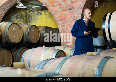 Mature serious winery worker wearing coat taking notes in wine cellar Stock Photo