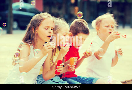 Four happy kids in school age sitting together and blowing soap bubbles Stock Photo