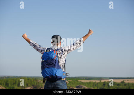 Photo back of tourist man with backpack ,hands up on hill in background of mountain expanses, blue sky during day Stock Photo