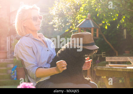 A woman relaxes in her garden with her dog on her lap who is wearing a hat Stock Photo