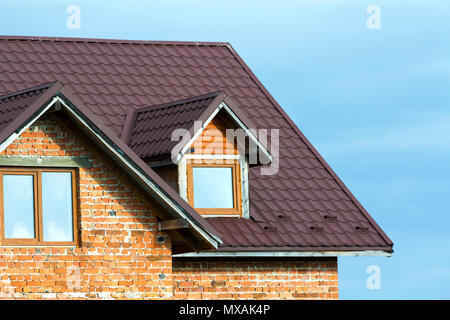 Close-up detail of new brick house top with brown shingle roof and plastic attic windows on bright blue sky background. Real estate property and profe Stock Photo