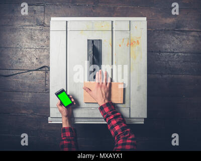 A young man operating a tablesaw is holding a chroma key smart phone and is being distracted from danger Stock Photo
