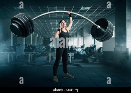 Female athlete with very big barbell talking by phone on workout in gim. Woman with weights, weightlifting sport concept Stock Photo