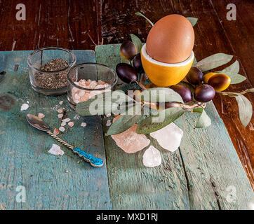 Single egg in a egg cup on a wooden surface with an assortment of crystal Hymalain pink salt and pepper. Olive decorative reef in the background. Stock Photo