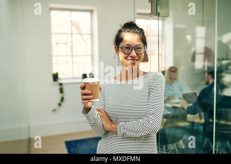 Smiling young Asian businesswoman wearing glasses holding a takeaway cup of coffee while standing in an office with colleagues at work in the backgrou Stock Photo