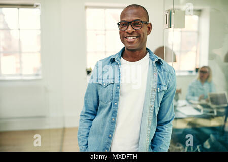 Casually dressed young African businessman wearing glasses and smiling while standing in an office with colleagues at work in the background Stock Photo