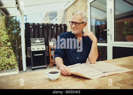 Senior man smiling contently while sitting at his patio table drinking a cup of coffee and reading the newspaper Stock Photo