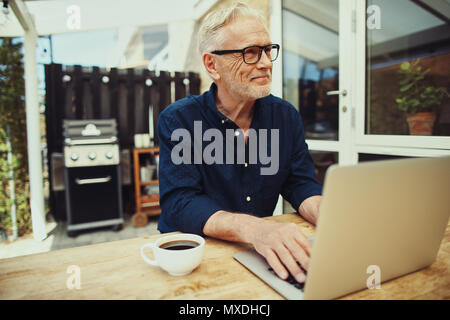 Smiling senior man sitting at his patio table outside working on a laptop and drinking a cup of coffee Stock Photo