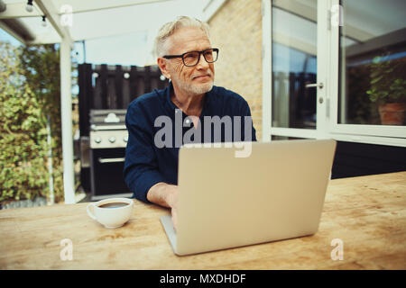 Smiling senior man sitting outside at his patio table drinking a cup of coffee and working with a laptop Stock Photo