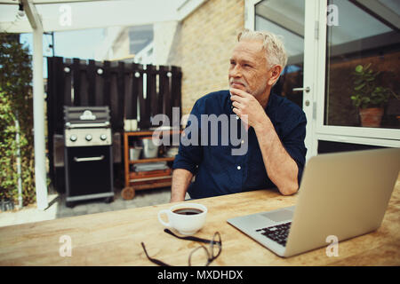 Smiling senior man sitting at a table outside on his patio thinking while using a laptop and drinking a cup of coffee Stock Photo