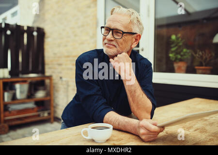 Smiling senior man sitting at a table outside on his patio thinking while using a tablet and drinking a cup of coffee Stock Photo