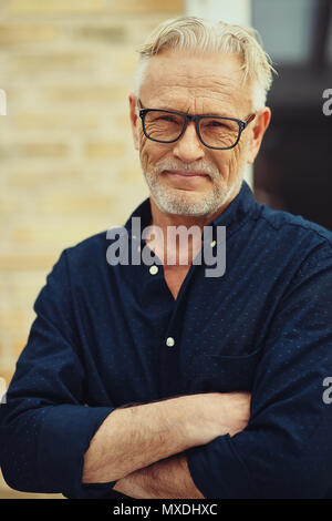 Smiling senior man with a beard and wearing glasses standing with his arms crossed outside in his yard at home Stock Photo