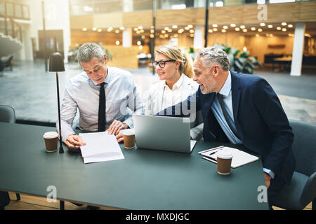 Three mature businesseople using a laptop and discussing paperwork while having a meeting together in the lobby of an office building Stock Photo