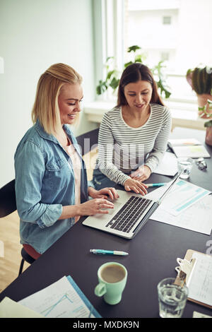 Two smiling young businesswomen talking together while sitting at a desk in an office working on a laptop Stock Photo