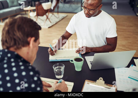 Focused African businessman sitting with a colleague at a table in an office writing notes and using a laptop Stock Photo