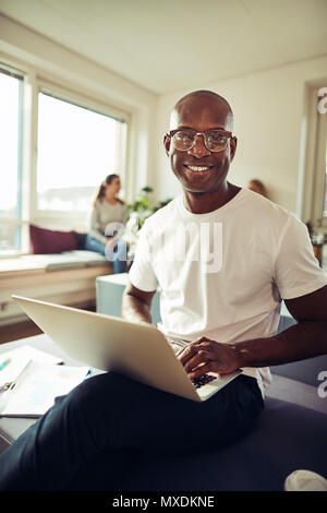 Smiling African businessman sitting at his desk in an office working on a laptop with colleagues sitting in the background Stock Photo