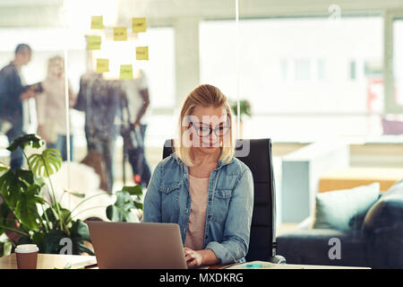 Casually dressed young businesswoman wearing glasses reading paperwork and working on a laptop while sitting at a desk in a modern office Stock Photo