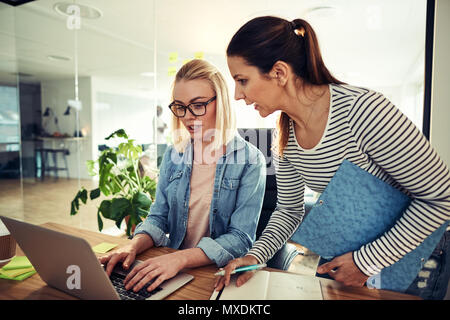 Two young business colleagues working together at a desk in an office discussing paperwork and using a laptop Stock Photo
