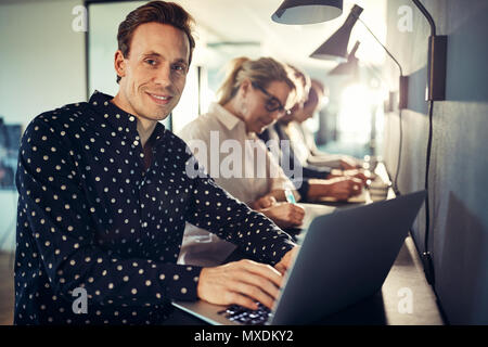 Smiling young designer working on a laptop while sitting in a row with coworkers at a table in a modern office Stock Photo