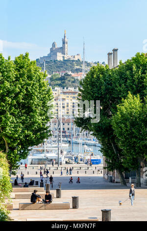 People strolling or resting on place Villeneuve-Bargemon and behind the trees, the Old Port of Marseille and Notre-Dame de la Garde basilica. Stock Photo