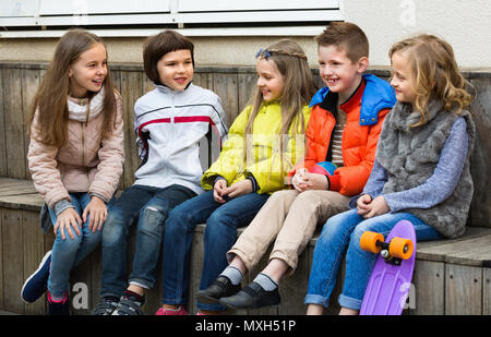 Group of  positive children sitting on bench and sharing secrets outdoors Stock Photo