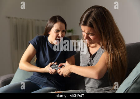 Two friends fighting for the tv remote control sitting on a couch in the living room at home Stock Photo