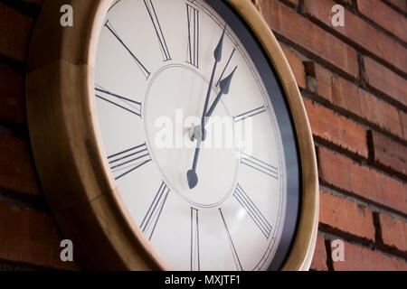 Side close–up of a wooden wall clock with roman numerals hanging in a red brick wall. Stock Photo