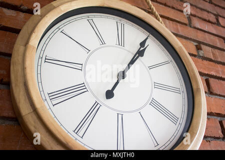 Close–up of a wooden wall clock with roman numerals hanging in a red brick wall. Stock Photo