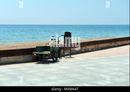 Green tricycle for adults parked next to green garbage can by harbor. Bicycle with luggage carrier. Stock Photo