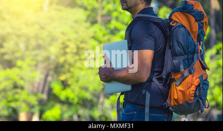 Man with a backpack standing in a park during day . Backpacker holding a laptop. Digital nomad concept Stock Photo