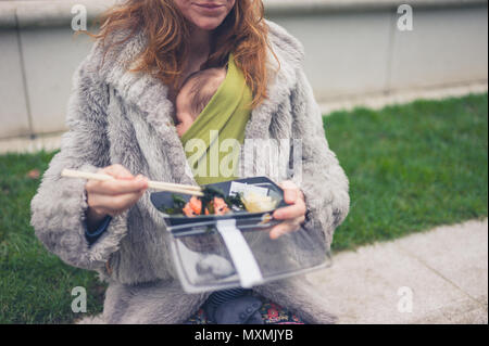 A young mother with a baby in a carrier sling is eating sushi outside Stock Photo