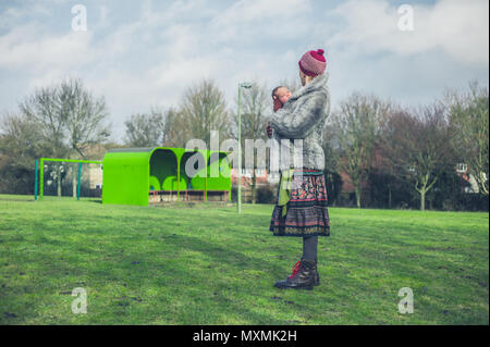 A young mother is standing on the grass in a park in winter with her baby in a carrier sling Stock Photo