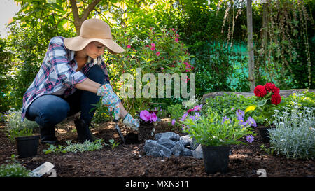 Beautiful female gardener wearing straw hat planting flowers in her garden. Gardening concept. Garden landscaping small business owner. Stock Photo
