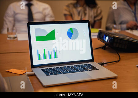 Front view of a computer plugged into a projector showing charts and graphs with information on. Unrecognisable people in the background. Stock Photo